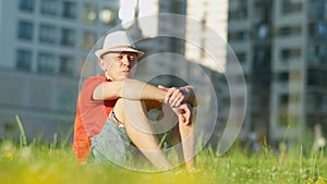 Man in hat is resting while sitting on the grass in a city park, tracking the camera