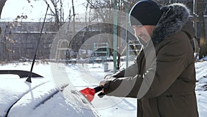 A man in a hat with a scraper in winter cleans the windshield or windshield of a car from snow and ice. Use a scraper or