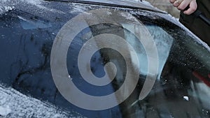 A man in a hat with a scraper in winter cleans the windshield or windshield of a car from snow and ice. Use a scraper or