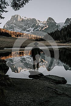 A man with a hat is standing by a mountain lake in the Dolomites. The mountains and the rest of the landscape are reflected in the