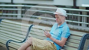 Man in hat uses mobile phone while sitting on city bench, camera tracking