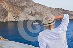 A man in a hat and white shirt is sitting with his back on the seashore, and staring at the yacht