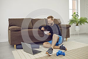 Man having break during fitness workout at home, eating healthy food and using laptop