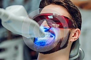 Man having teeth whitening by dental UV whitening device,dental assistant taking care of patient,eyes protected with glasses.