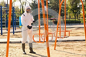 Man in hazmat suit with disinfectant sprayer near swing at children`s playground. Surface treatment during coronavirus pandemic