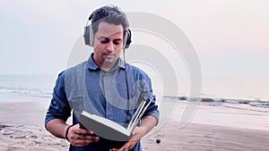 Man with headphones reading a book and walking on a tranquil beach at dusk