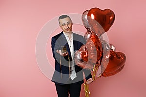 A man with heart balloons on a pink background holds out a box with a wedding ring. Betrothed on Valentine`s Day, man proposing,