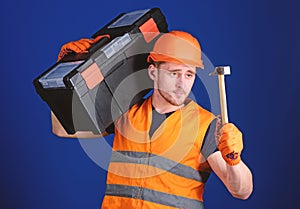 Man in helmet, hard hat carries toolbox and holds hammer, blue background. Handyman concept. Worker, repairer, repairman