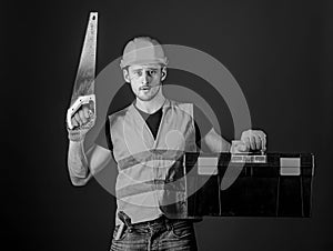 Man in helmet, hard hat carries toolbox and holds handsaw, blue background. Carpenter concept. Worker, repairer