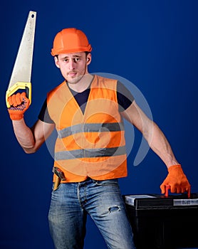 Man in helmet, hard hat carries toolbox and holds handsaw, blue background. Worker, repairer, repairman on serious face