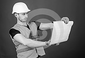 Man in helmet, hard hat holds building plan, controls works, red background. Engineer, architect, builder on strict face