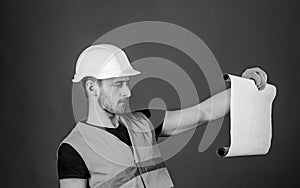 Man in helmet, hard hat holds building plan, controls works, red background. Engineer, architect, builder on strict face