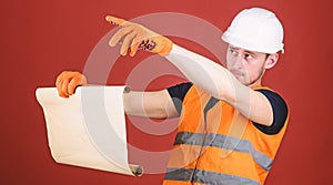 Man in helmet, hard hat holds building plan, controls works, red background. Foreman concept. Engineer, architect