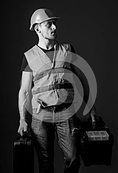 Man in helmet, hard hat holds toolbox and suitcase with tools, blue background. Worker, handyman, repairman, builder on