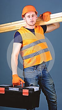 Man in helmet, hard hat holds toolbox and wooden beams, grey background. Carpenter, woodworker, labourer, builder on
