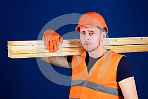 Man in helmet, hard hat and protective gloves holds wooden beam, blue background. Wooden materials concept. Carpenter