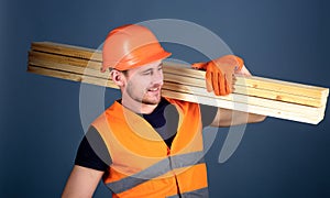 Man in helmet, hard hat and protective gloves holds wooden beam, grey background. Carpenter, woodworker, labourer