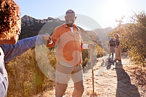 Man Helping Woman On Trail As Group Of Senior Friends Go Hiking In Countryside Together