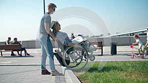 A man helps a girl in a wheelchair to go down from the curb to the asphalt.  A man is pushing a wheelchair