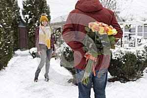 man hides a large bouquet of roses behind his back, and a happy woman stands in front of him in the background