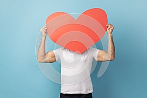 Man hiding behind big red paper heart, holding symbol of love care, cardiac issues.