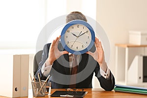 Man hiding face behind clock at table in office. Time management concept