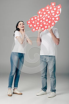 Man hiding face behind red paper cut cards with hearts symbol near excited girl on grey background