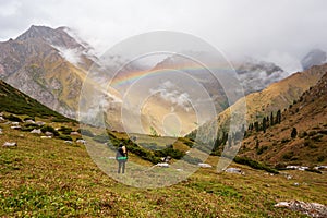 Man hiker with backpack looking at rainbow. Male Backpacker in rainy mountains Karakol valley on trail to Ala Kul lake. Karakol