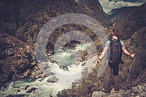Man hiker with backpack standing on the edge of the cliff with epic wild mountain river view.