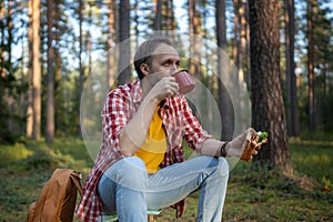 Man hiker drinking tea from mug, eating sandwich during hiking in forest resting stopped for break