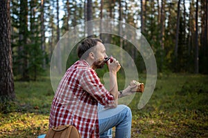 Man hiker drinking tea from mug, eating sandwich during hiking in forest resting stopped for break