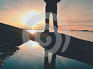 Man hiker legs in high boots mirrored in water pool, sea with sunset sun. Tourist figure on rocky