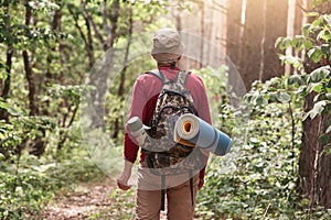 Muž s turistickým vybavením na procházce v letním lese, na sobě ležérní oblečení, pózuje pozpátku, drží batoh a karimatku