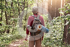 Man with hiking equipment walking in summer forest, wearing casual clothing, posing backwards, holding backpack and sleeping mat