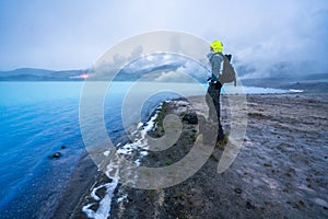 Man with hiking gear stands on shore looking towards Geothermal powerplant at the blue lagoon in Jarabodin/Myvatn