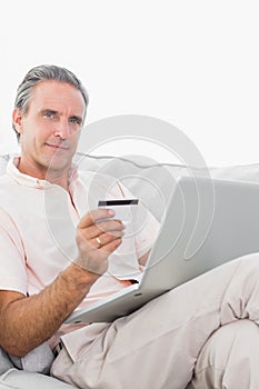 Man on his couch using laptop for shopping online smiling at cam