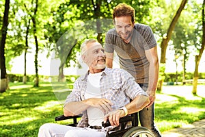 A man and his elderly father are walking in the park. A man carries his father in a wheelchair