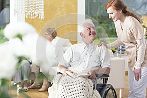 A man in his old age ia a wheelchair talking to a personal assistant in a common room of a rehabilitation center.