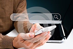 The man is on his phone while works on a laptop computer. With a blank search bar, the concept is Search engine Browsing Internet.