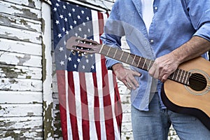 Man holding acoustic guitar in front of abandoned barn with American Flag