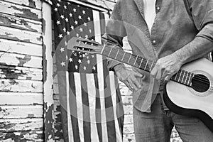 Man holding acoustic guitar in front of abandoned barn with American Flag