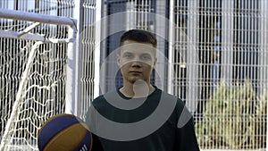 Man holding a basketball ball near the metal fence of a sports ground. Media. Young man at outdoors basketball field.