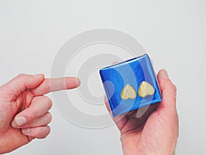 Man holding a blue gift box with two golden glitter hearts in one hand pointing with other. White background. Romance and love