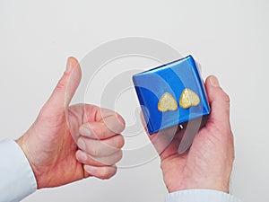 Man holding a blue gift box with two golden glitter hearts in one hand thumb up with other. White background. Romance and love
