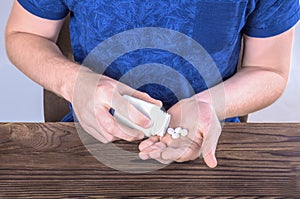 A man holding a bottle full of medicine. A young male pouring drugs out of a white bottle. An ill guy on a gray background.