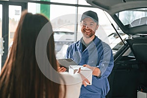 Man is holding box and notepad. Young woman is visiting auto mechanic in garage. Repair service