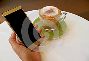 Man holding cellphone in his hand beside a cup of hot cappuccino coffee on the white table