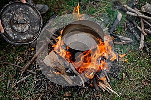 Man holding a cover near the pan with water standing on the bonfire