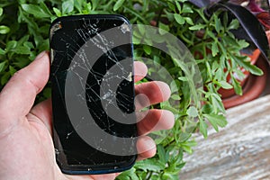 Man is holding a crashed black smartphone in hand on the green leaves plant background. Broken lcd touch screen. Information