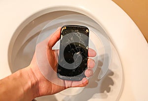 Man is holding a crashed black smartphone in hand over the toilet bowl. Broken lcd touch screen in the bathroom. Information